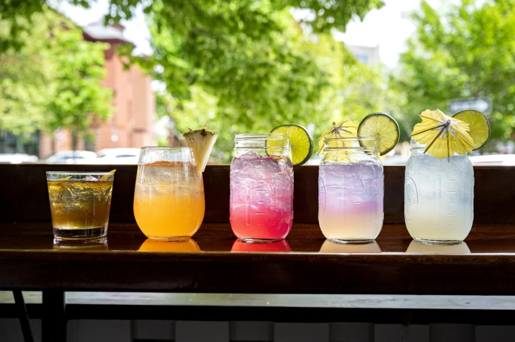 Five colorful cocktails in mason jars and glasses garnished with citrus and paper umbrellas sit on an outdoor wooden ledge.