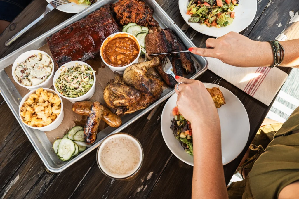 An overhead view of a person using a fork and knife to slice into barbequed meats from a large metal platter with sides.