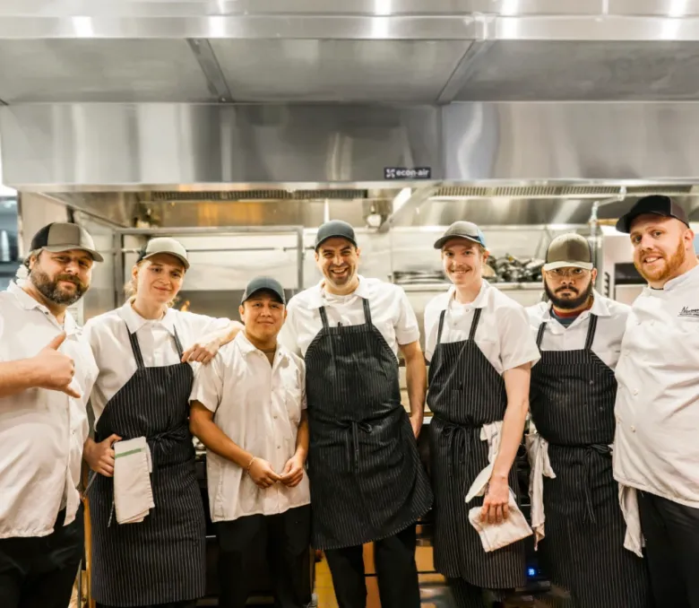 Seven kitchen staff members in white shirts and black aprons smile together in a commercial kitchen.
