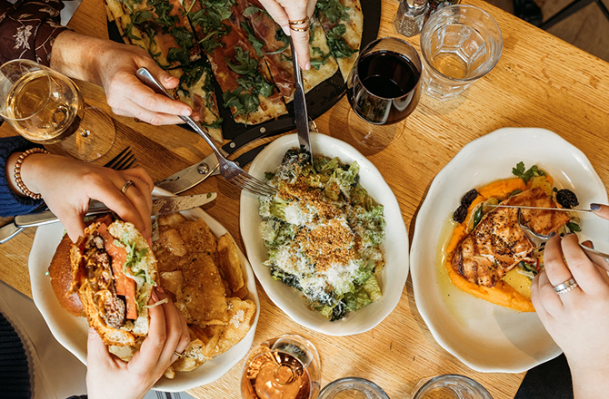 A dining table shared by friends, featuring dishes including a burger, salad, grilled fish with squash puree, and glasses of wine.