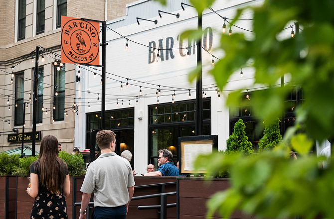 Exterior of Bar 'Cino with an outdoor patio setup, overhead string lights, and people walking by.