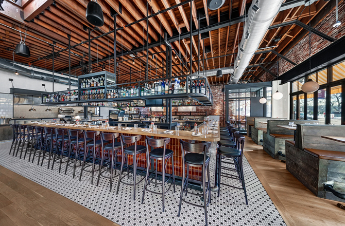 Modern restaurant interior featuring a long wooden bar with tall stools, exposed wood beam ceilings, and patterned tile flooring.