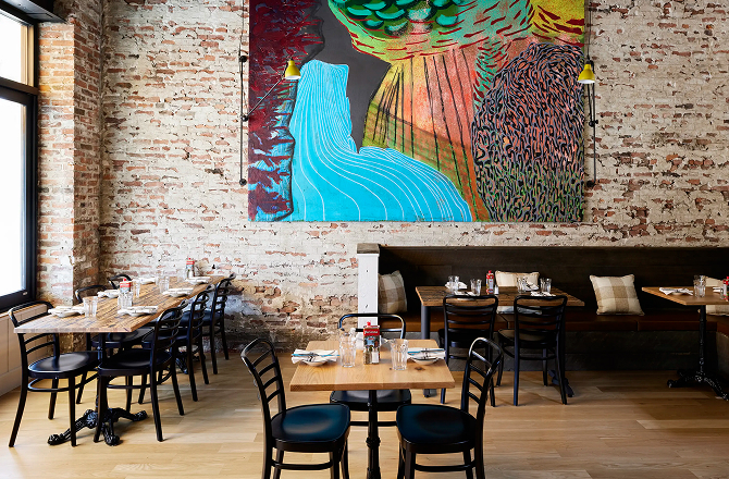 Spacious restaurant dining area featuring wooden tables, black chairs, and a colorful tapestry on an exposed brick wall.