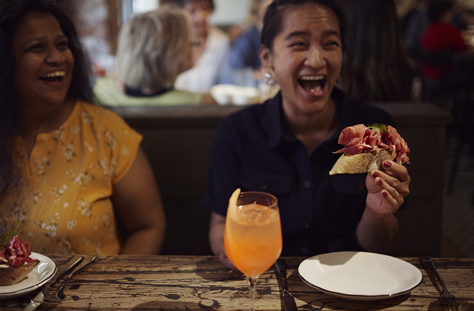 Two women laughing while sitting at a table with sandwiches, plates, and a cocktail.