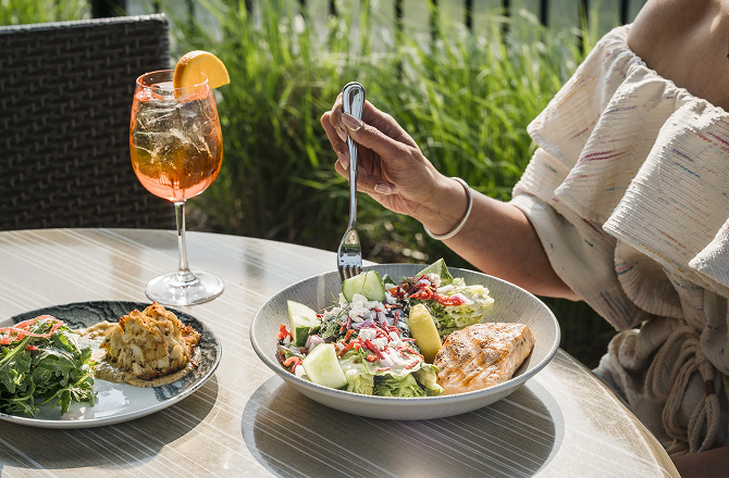 Person dining outdoors at a table with plated salads, a crab cake, and an orange spritzer cocktail.