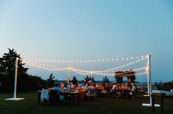 Outdoor evening event lit by string globe lights on a lawn overlooking the ocean.