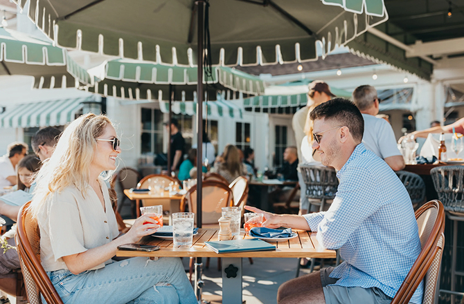 Young couple dining outside at a patio table on a sunny day under a striped umbrella.