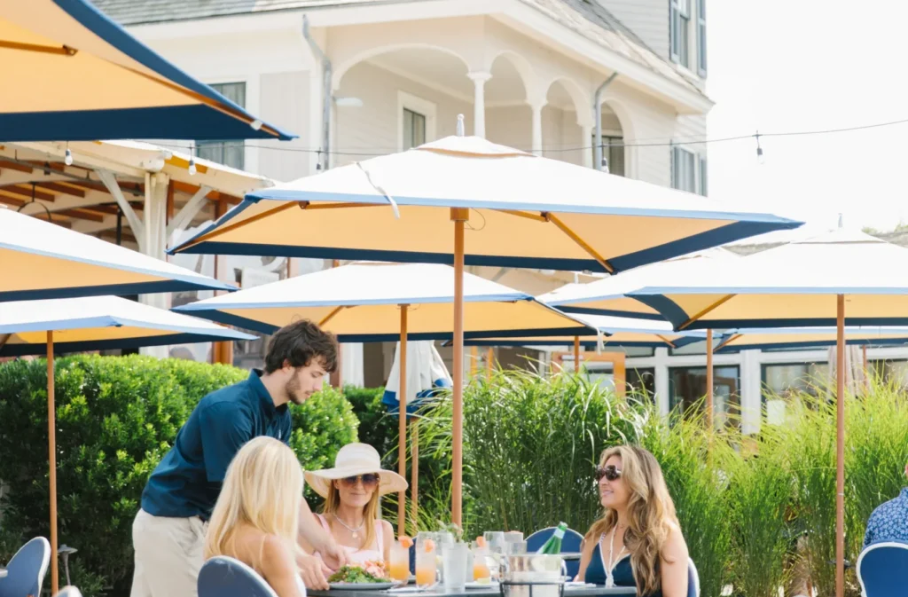 A waiter serves food to women seated at an outdoor patio table under large umbrellas in front of a white building.
