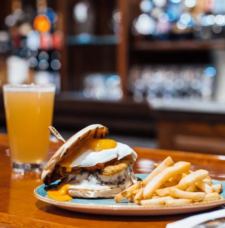 A cheeseburger topped with a fried egg served alongside French fries and a glass of beer on a wooden bar top.