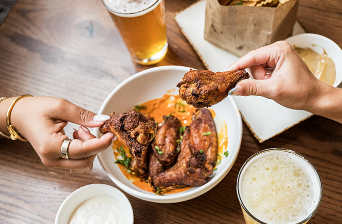 Two diners reach for glazed chicken wings in a white bowl paired with draft beer and potato chips.