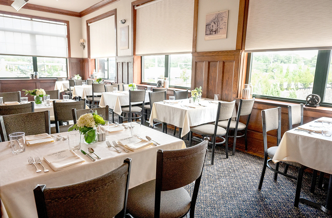 Restaurant dining room set with white tablecloths, wooden chairs, floral centerpieces, and large sunlit windows.