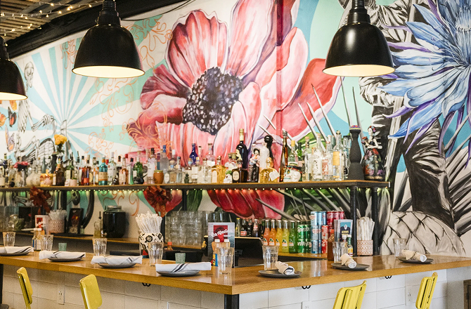 Vibrant bar area featuring yellow chairs, a wooden counter, black pendant lights, and a colorful floral mural.