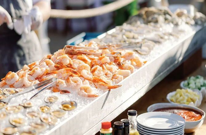 Raw seafood bar station displaying chilled cooked shrimp and oysters on ice with serving tongs.