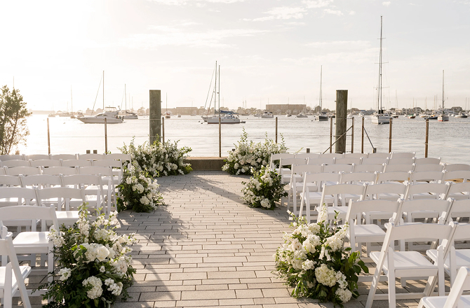 Outdoor wedding ceremony setup on a paved patio with white chairs, floral arrangements, and sailboats in the harbor.