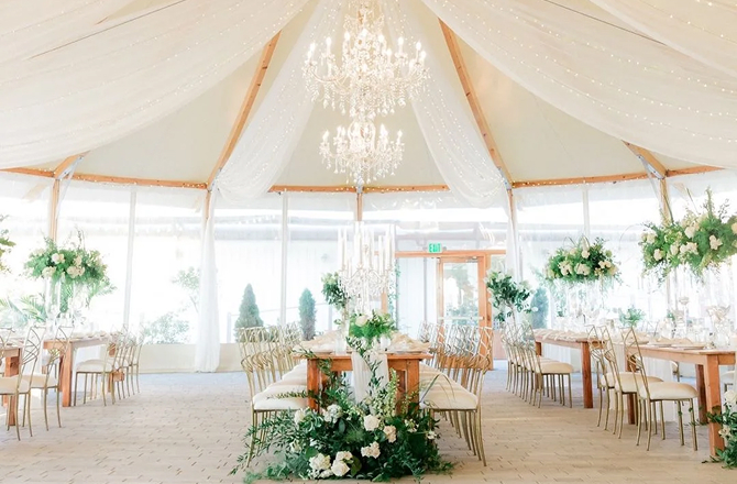 Elegant white event tent filled with set dining tables, crystal chandeliers, and lush floral greenery arrangements.