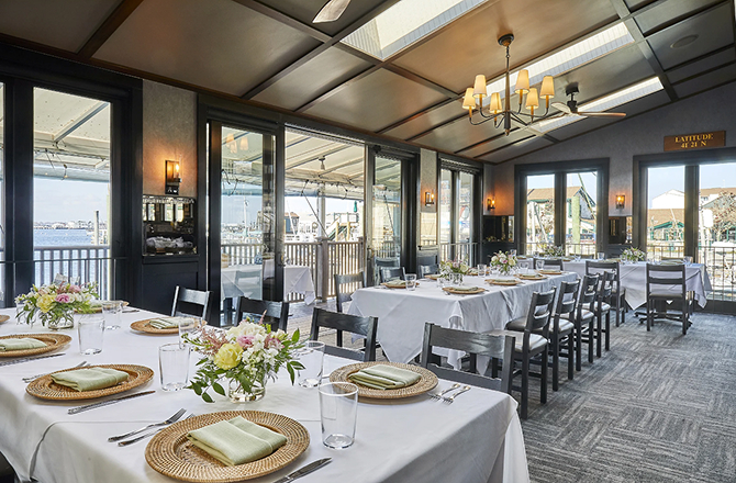 Sunlit restaurant dining room with long tables set with white tablecloths and floral arrangements.