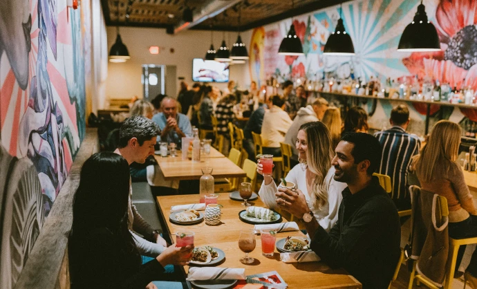 A lively restaurant dining area filled with guests seated at wooden tables enjoying drinks and meals beneath a large colorful mural on the wall.
