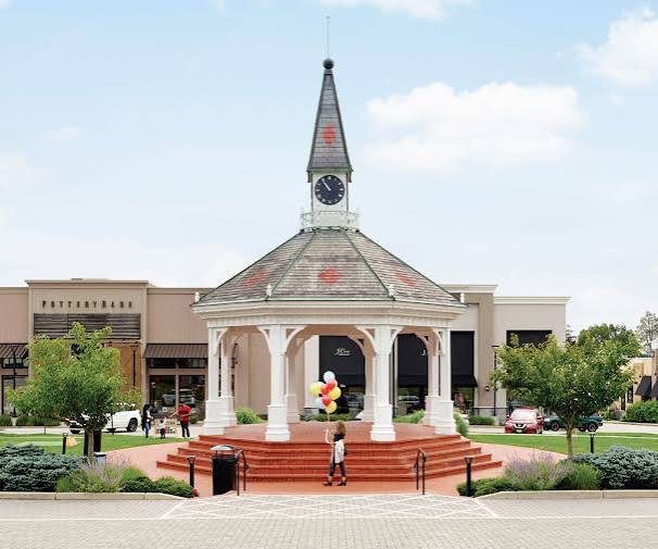 A large white gazebo with a clock tower stands in an outdoor shopping center plaza with storefronts and trees in the background.