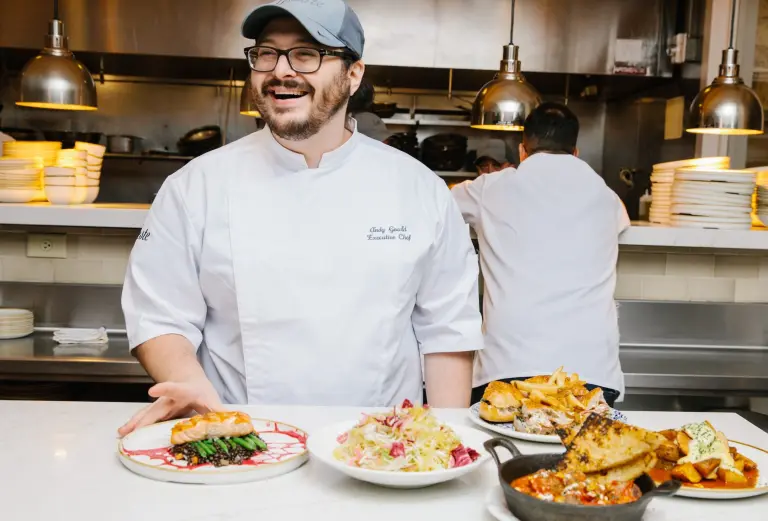 A smiling chef in a white coat stands in a kitchen behind a counter displaying multiple plates of prepared food.