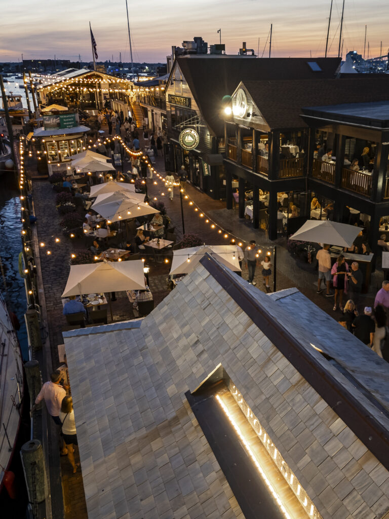 Elevated view of a bustling harborside boardwalk at dusk with outdoor dining under string lights and umbrellas.